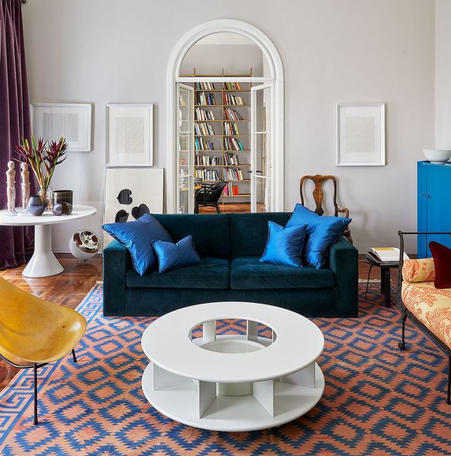 high ceiling white painted apartment with inset niche ceiling and a arched doorway with glass doors and transept and at center is a dark blue green velvet sofa with paul newman blue silk throw pillows on it sitting on a pink carpet with blue cut out triangle shapes and a white cocktail table in front with a cutout at center and a white pedestal table at the back and a mustard armless char in the left foreground