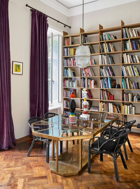 dining room with a wall bookcase behind it and an exaggerated octagonal glass table with gold frame and simple black chairs pulled up to it on a chevron pattern wood floor and hanging off the window to the left are the ripe plum colored draperies puddling on the floor