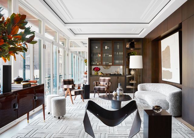 living room long shot with a white square design rug spanning the length of it and a rounded sofa against the right wall which is dark wood paneling and some cabinetry at the far end some behind glass and on the right a series of tall doors that open to the view