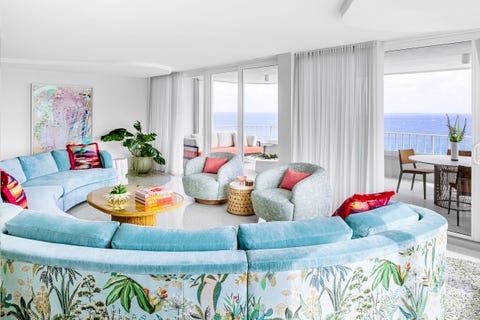 Fawn Galli - Elle Decor half moon sofa in a green velvet and a flowered print on the back and faces floor to ceiling windows leading to a terrace with a round table overlooking the water a rattan coffee table is in the center