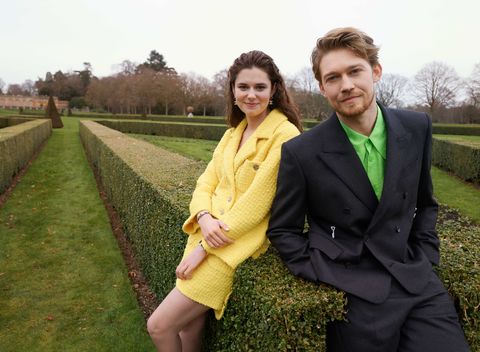 alison oliver and joe alwyn lean against a hedge