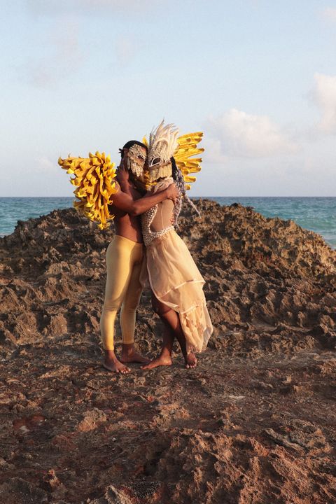 a still from 'ebbó﻿' shows one a person in gold leggings wearing wings and a mask embracing a person with a feathered mask wearing a beaded and diaphanous gown, standing in sand in front of the water