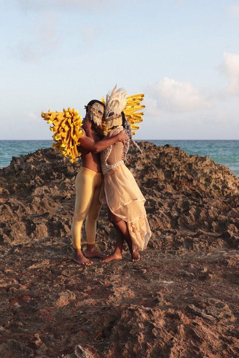 a still from 'ebbó﻿' shows one a person in gold leggings wearing wings and a mask embracing a person with a feathered mask wearing a beaded and diaphanous gown, standing in sand in front of the water