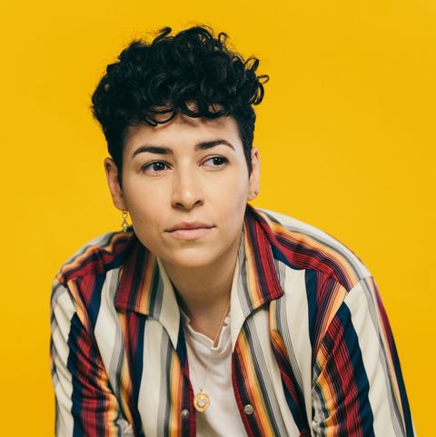 a photo of denice frohman shows the poet with dark, short curly hair, wearing one earring, a white t shirt and an unbuttoned short sleeve shirt with white pants