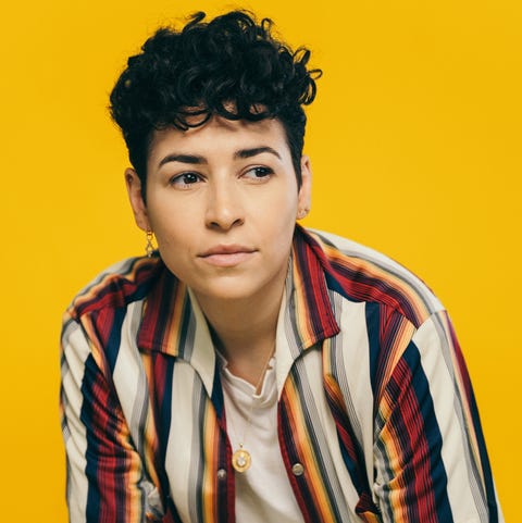 a photo of denice frohman shows the poet with dark, short curly hair, wearing one earring, a white t shirt and an unbuttoned short sleeve shirt with white pants