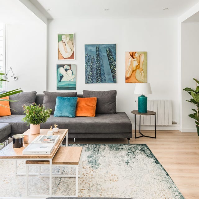 Salón abierto con sofá en L gris con cojines de colores, paredes blancas decoradas con cuadros y una palmera salón abierto con sofá en l gris con cojines de colores, paredes blancas decoradas con cuadros y una palmera