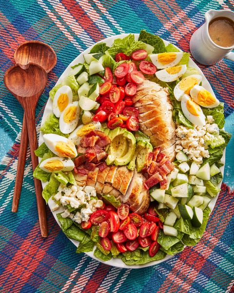 cobb salad in an oval white serving bowl with two wooden serving utensils and a small container of dressing to the side