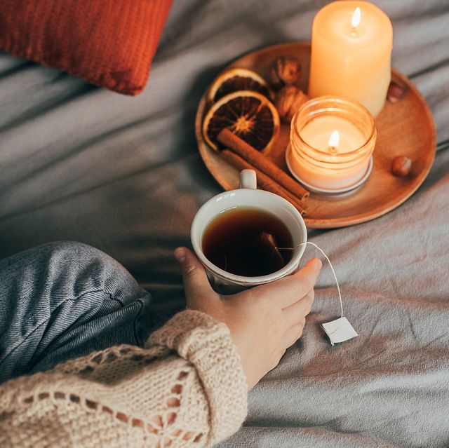 person holding tea in bed with candles, cinnamon sticks and dried oranges