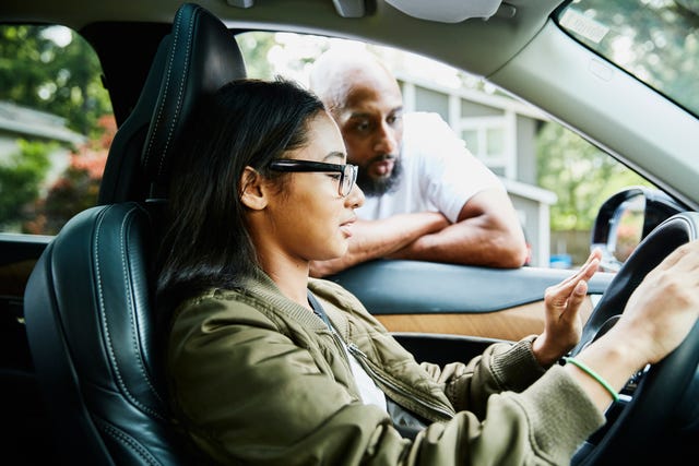 father leaning through window of car while teaching daughter to drive