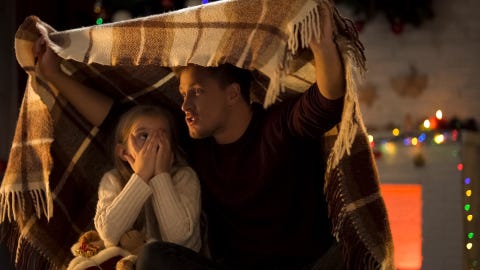 dad telling scary story for little girl covering her face with her hands and sitting under plaid blanket