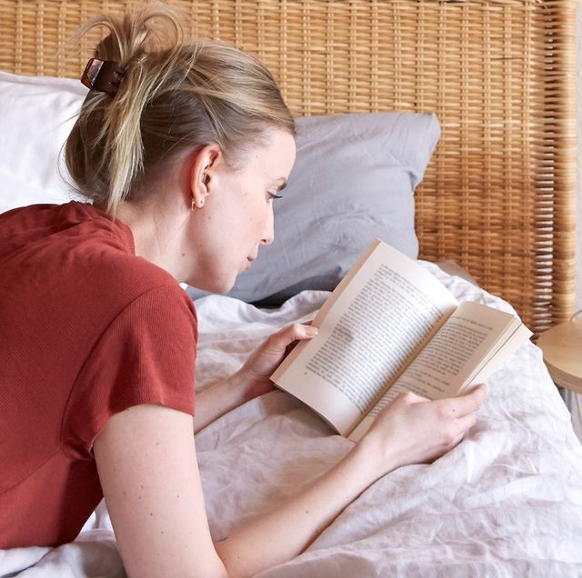 woman wearing a red shirt and jeans lying on bed reading a feel good book in sunlight