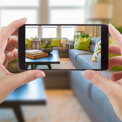 female hands holding smart phone displaying photo of house interior living room behind