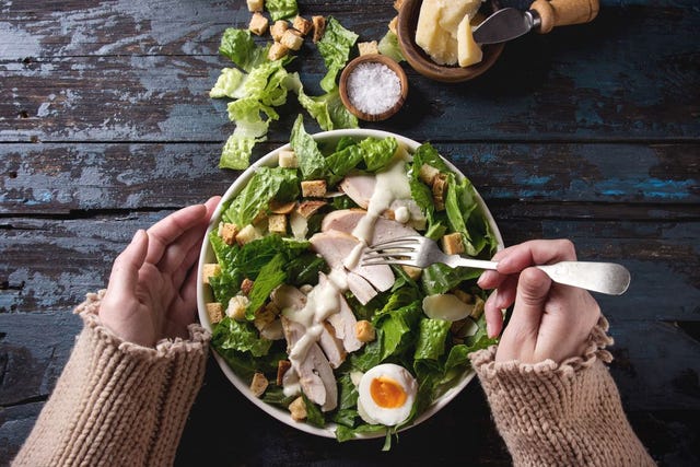 female hands take by fork classic caesar salad with chicken breast in white ceramic plate served with ingredients above over old dark blue wooden background flat lay rustic style