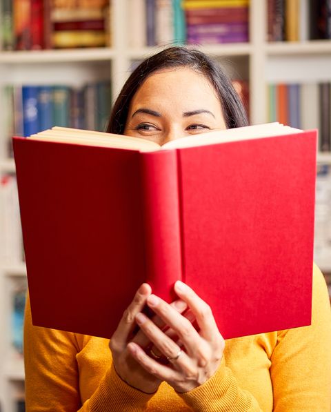 female young behind book with face covered for a red book while smiling