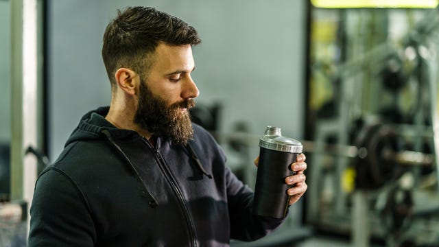 Front view portrait of a young Caucasian male athlete in a black hoodie standing in the gym holds a protein supplement shaker supplement when training a copy space drink with black hair and beard