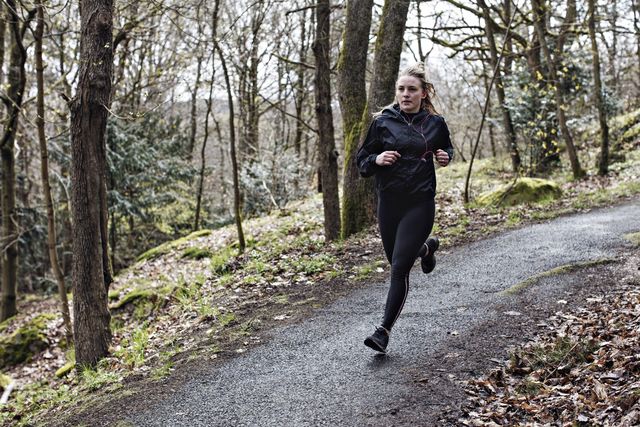 full length determined female athlete running on narrow road in forest