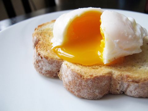 a single poached egg on artisan hand sliced toasted bread the egg is split to reveal a runny yolk white dinner plate, non descript dark wood table and chair just about visible in the background