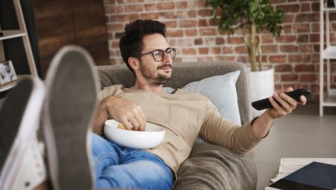 Content man lying on couch at home with bowl of potato chips watching TV