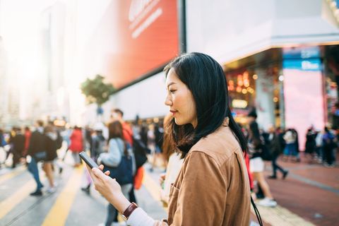 Young woman checking on mobile phone while crossing street and commuting in busy downtown city street
