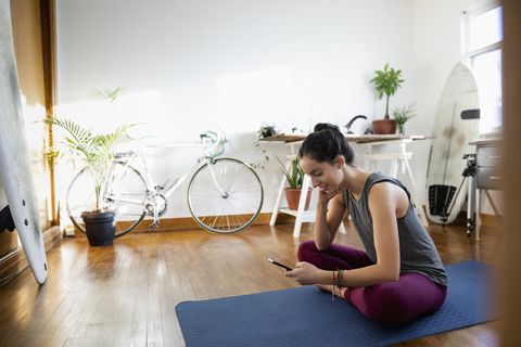 Young Latinx woman with smart phone on yoga mat in apartment