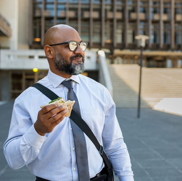 mature african businessman in formal clothing eating brown bread sandwich outdoor senior entrepreneur wearing spectacles standing outside office building, looking away while enjoying vegetable sandwich business man having lunch on the move