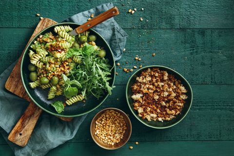 summer vegetarian pasta salad with broccoli pesto, peas, arugula, olives, pine nuts and bread crumbs on dark green background top view