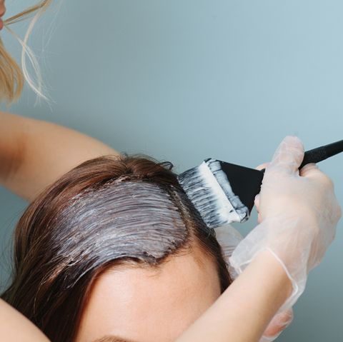 focused blonde female hairdresser in apron is applying white developer on clients hair over blue background close up