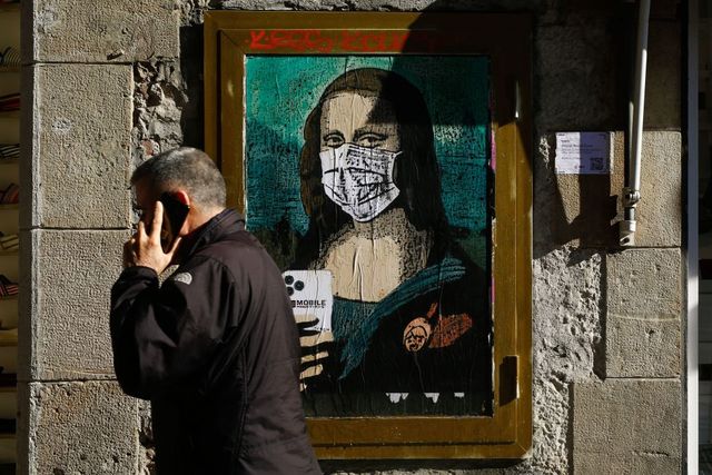 a man holding his mobile phone walks past a poster by italian urban artist salvatore benintende aka tvboy depecting leonardo da vincis mona lisa  wearing a protective facemask and holding a mobile phone reading mobile world virus in a street of barcelona on february 18, 2020, a week after the world mobile congress was cancelled due to fears stemming from the coronavirus that sparked an exodus of industry heavyweights photo by pau barrena  afp  restricted to editorial use   mandatory mention of the artist upon publication   to illustrate the event as specified in the caption photo by pau barrenaafp via getty images