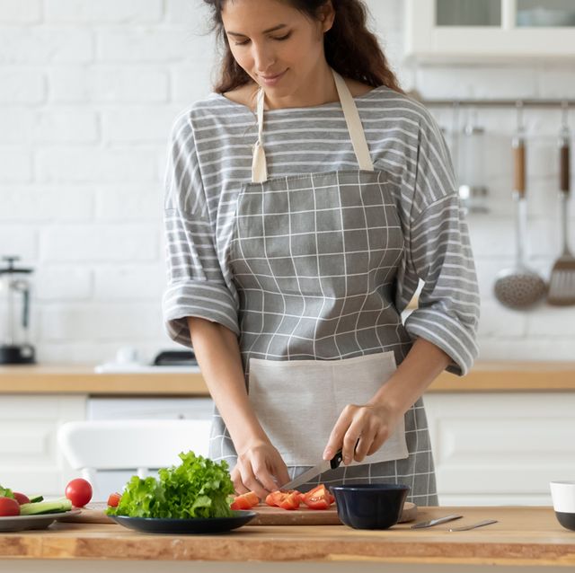 millennial woman in apron stand at kitchen table chop vegetables preparing salad for dinner or lunch, young housewife cooking healthy tasty breakfast at home, dieting, vegetarian concept