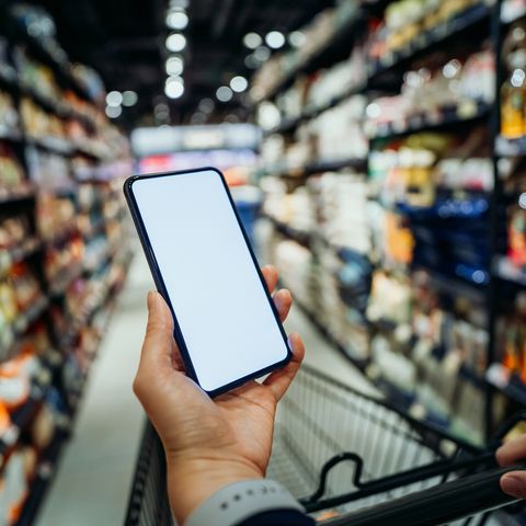close up of womans hand with trolley using smartphone and checking shopping list while grocery shopping in a supermarket