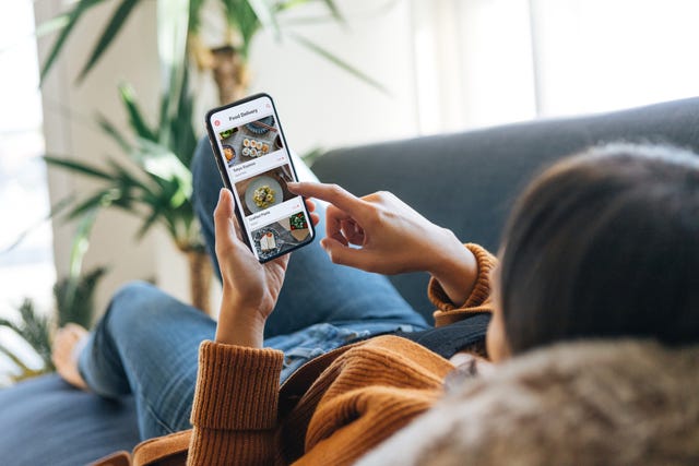 Au-Dessus De L'épaule D'une Jeune Femme Choisissant La Nourriture Dans Le Menu Sur L'application Mobile En Position Couchée Sur Le Canapé À La Maison