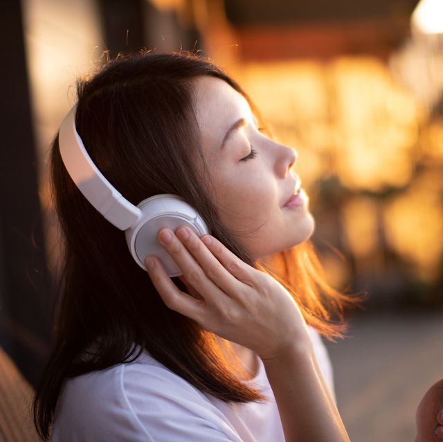 portrait of smiling young asian woman with eyes closed enjoying music over headphones and using smartphone, relaxing on the balcony against sunlight