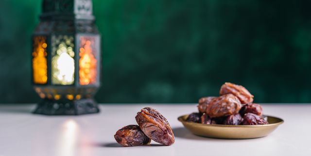 dates close up in the foreground on the distant plan a slightly blurry burning, lighting, glowing ramadan lantern on a white table, textured dark green wall background place for text on the right