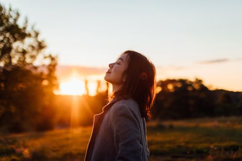 side portrait of young asian woman with eyes closed inhaling fresh air, against sunset in the sky