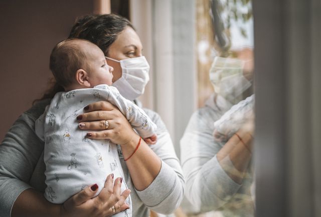 mother with protective face mask holding her newborn baby and they looking trough the window