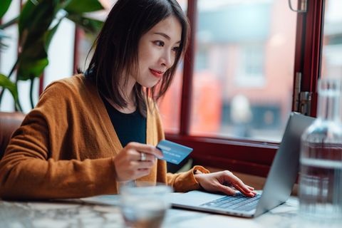 smiling young asian woman shopping online with laptop, making mobile payment with credit card, sitting near the window in a cafe online payment concept shopping online makes life easier