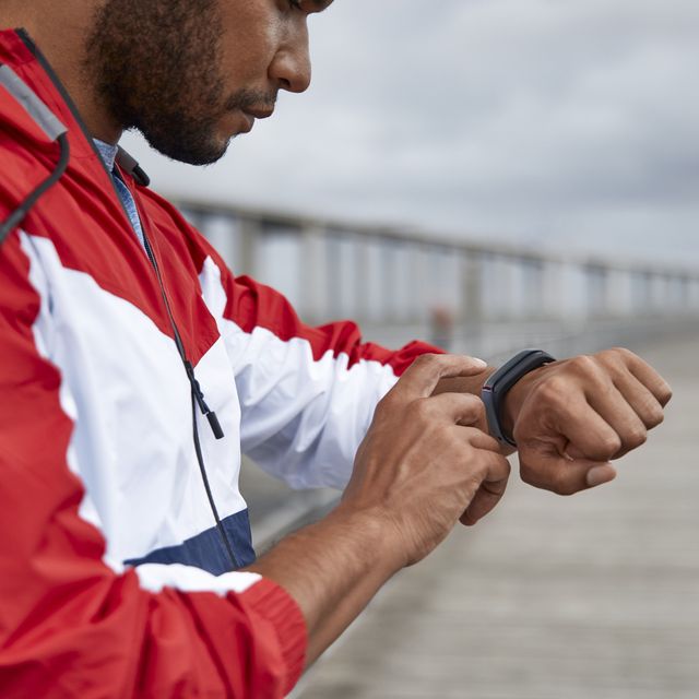 african american young man with earphones wearing wind jacket tapping fitness tracker by river bridge