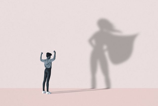 young woman flexing muscles in front of large superhero shadow on pink background