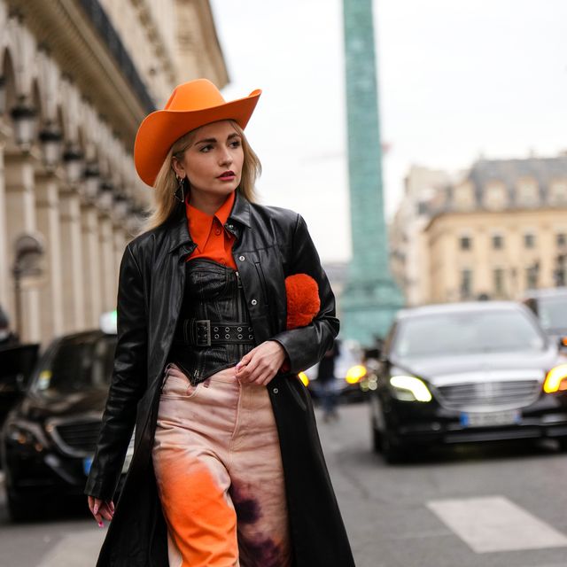 paris, france   march 01 a guest wears an orange felt  wool cow boy hat, a silver and black pendant earrings from prada, gold earrings, an orange shirt, a black shiny leather zipper  studded  shoulder off corset, a black shiny leather long coat, an orange fluffy handbag, pale orange and orange tie and dye denim large jeans pants, black shiny leather block heels ankle boots, an orange large ring , outside koche, during paris fashion week   womenswear fw 2022 2023, on march 01, 2022 in paris, france photo by edward berthelotgetty images