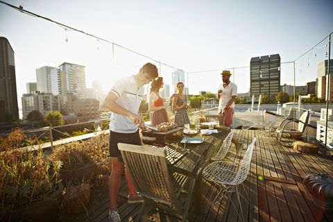 group of friends standing around table set for dinner on rooftop deck man checking barbecue at sunset