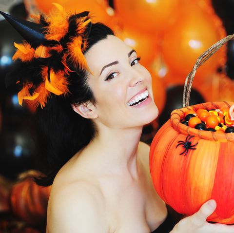 beautiful smiling brunette woman dressed up for halloween with a feather hat, holding an orange basket filled with candies, with black and orange balloons in the background
