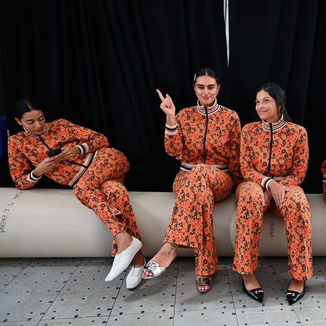 Models at the historic Birch SS23 in matching orange dresses sit together chatting and looking at their phones.