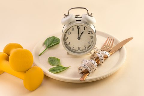 healthy food composition with cutlery, plate, measuring tape, yellow dumbbell, green vegetable salad leaves and alarm clock on pastel beige background dieting and keep fit close up copy space front view