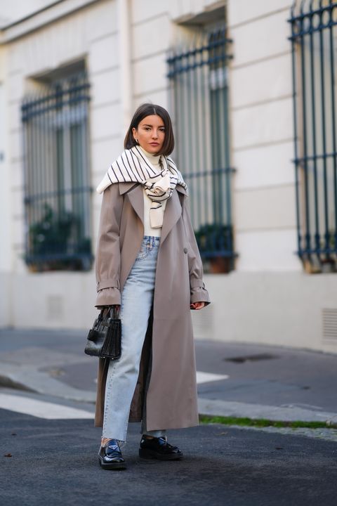 paris, france   october 19 alexandra pereira wears silver earrings, a white ribbed wool turtleneck pullover, a white with navy blue striped pullover tied on the shoulders, a pale gray long belted trench coat, high waist blue faded denim large jeans pants, a black shiny leather crocodile print pattern sac du jour handbag from saint laurent, black shiny leather loafers from prada, during a street style fashion photo session, on october 19, 2021 in paris, france photo by edward berthelotgetty images