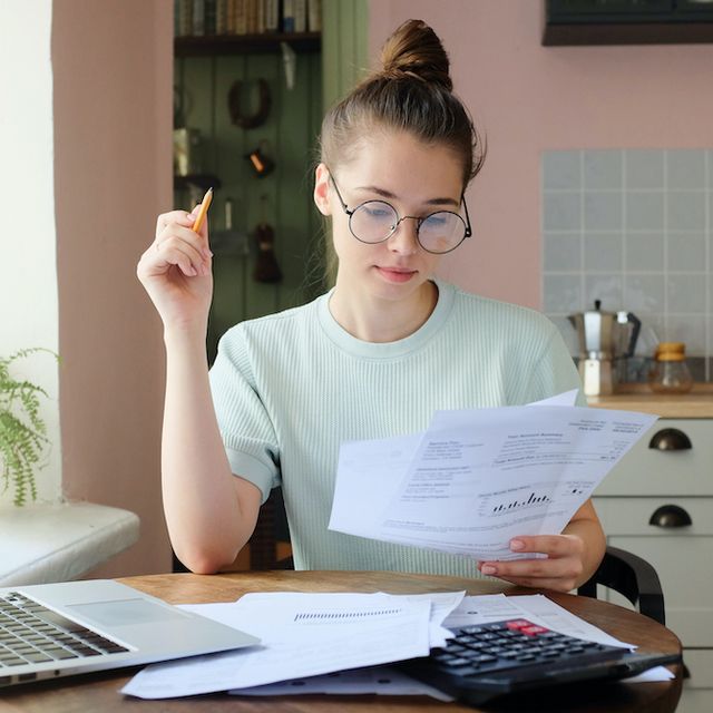 indoor picture of young european female sitting at home at table reading data in sheets of paper with pencil in order to figure out something important and check it, feeling relaxed and confident