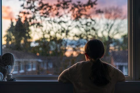 Rear view shot of a young girl looking out of her window at sunset a young girl, with a ponytail, looks out of her bedroom window as the sun is setting the view through the window is blurry but shows a residential area