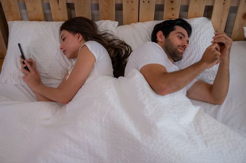 young couple in bed late at morning using smart phones obsessed with games, social media and apps ignoring each other in relationship communication problems and internet mobile addiction concept