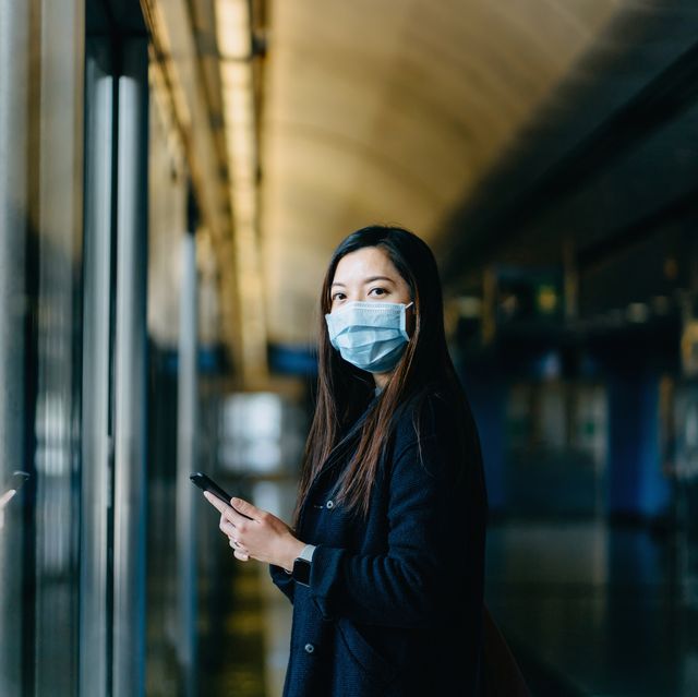 asian woman with protective face mask using smartphone while commuting in the city and waiting for subway in platform