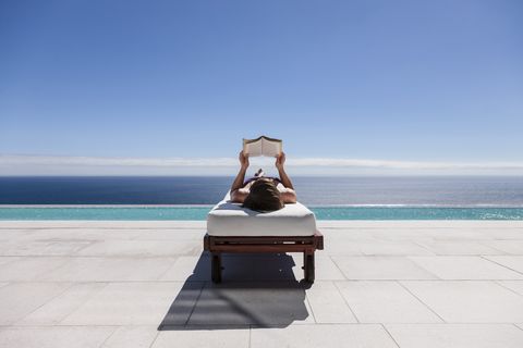 Woman reading on lounge chair at poolside overlooking ocean