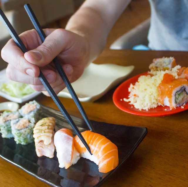 a close up photo of a man eating salmon sushi roll theres a set variation sushi in front of the man on the left side, theres a wasabi and soy sauce at the top right picture, there is an ice green tea the man holding a chopstick to eat a sushi roll